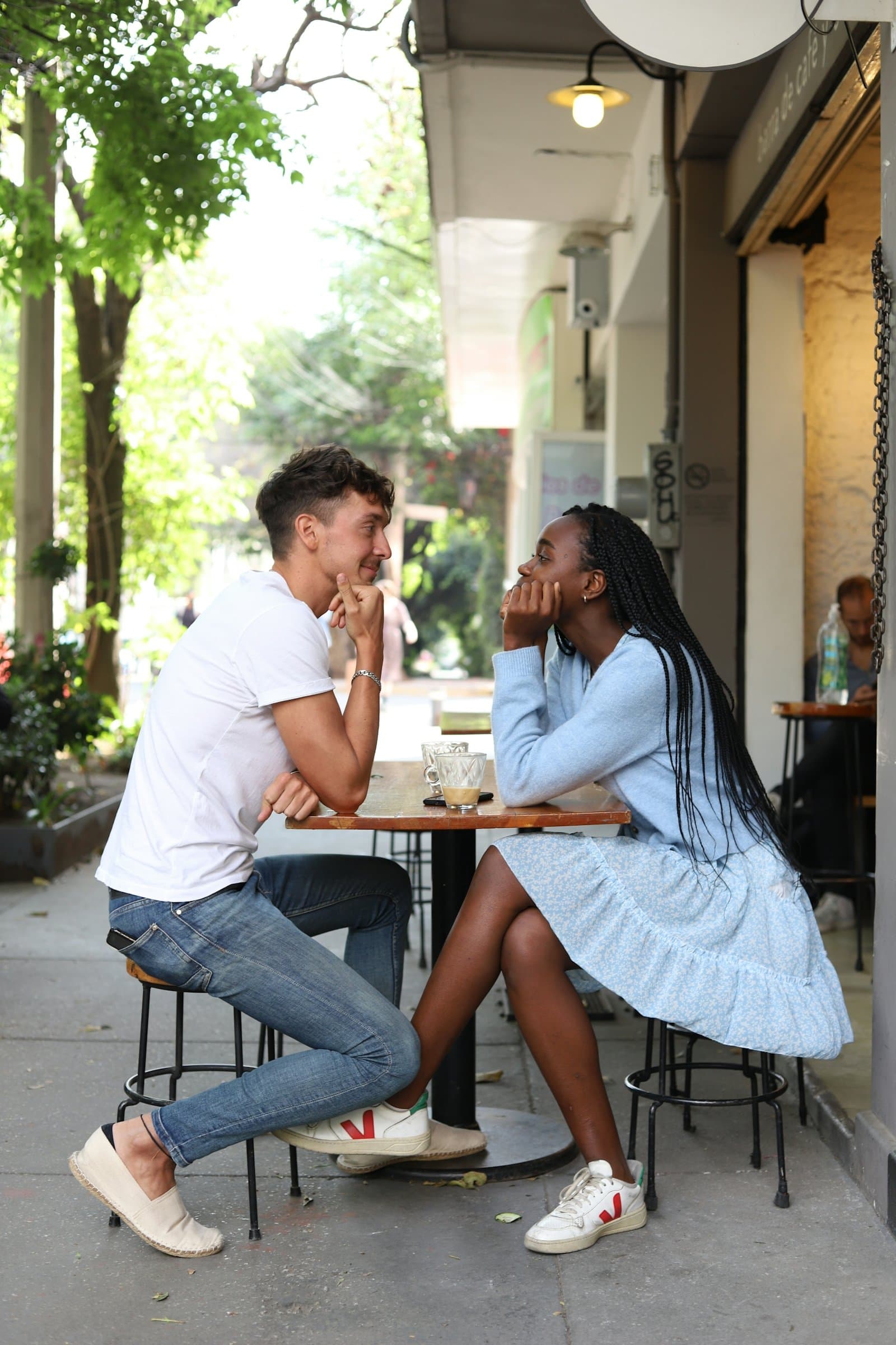 A young man and woman at a small footpath café table, chins in their hands, smiling at each other