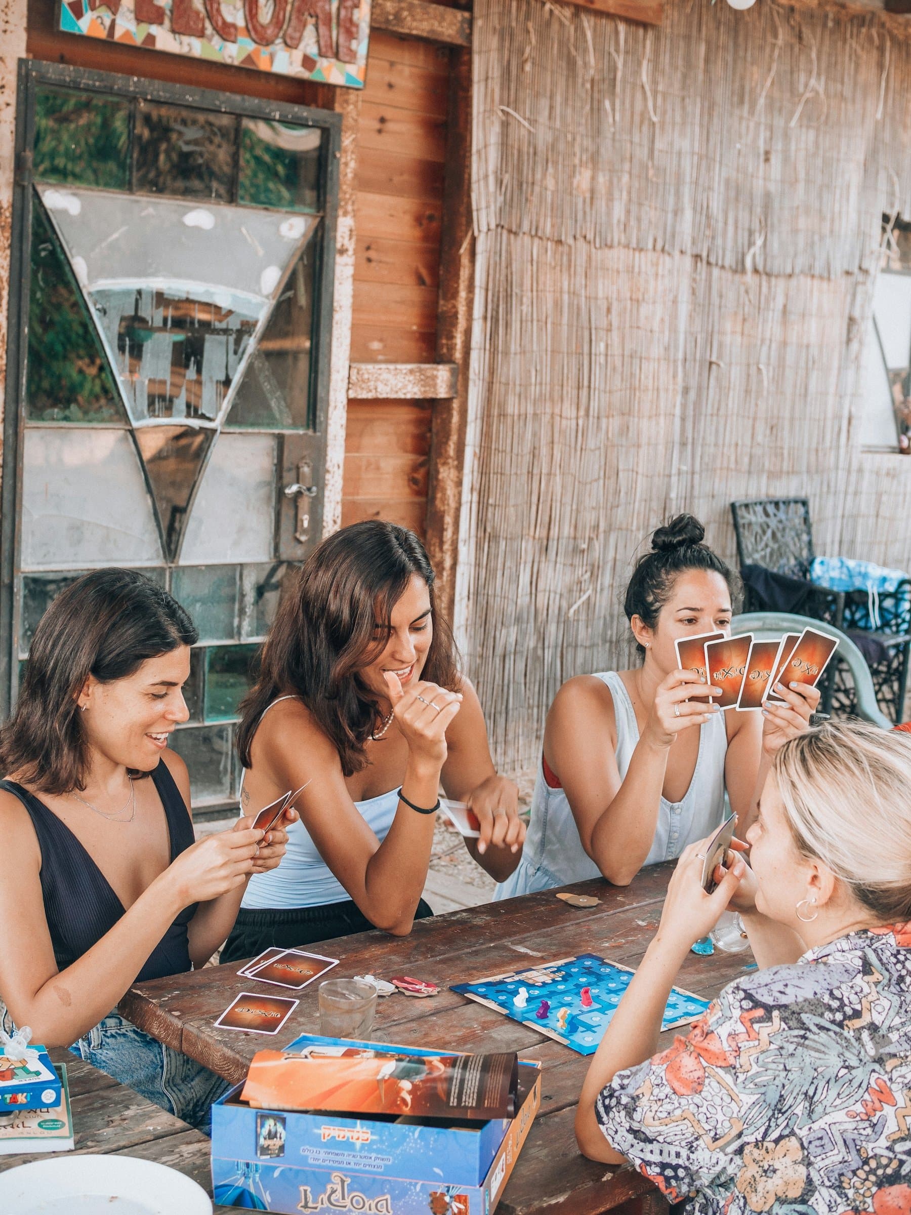 Four women laughing over a card game at a weathered wooden table outside a rustic café