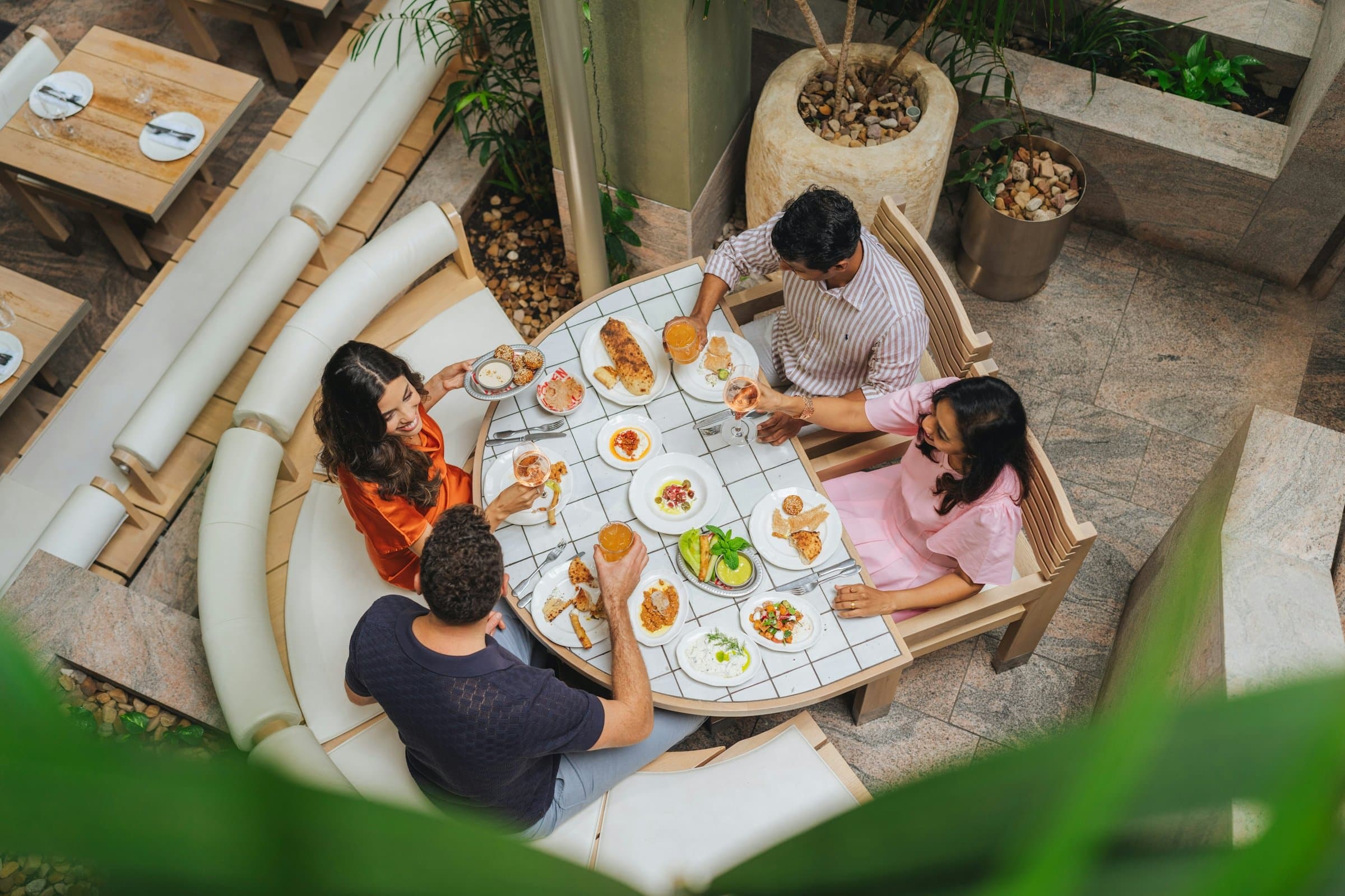 Four friends sharing small plates around a table in a leafy courtyard restaurant, seen from above