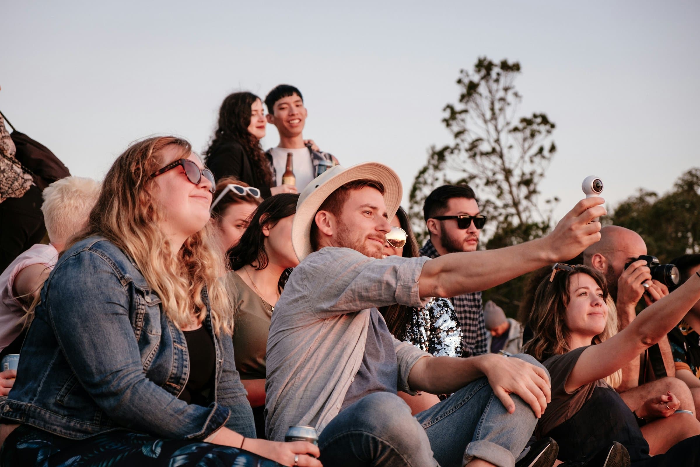 A group of friends sitting together on a hilltop at golden hour, one holding up a small camera for a group shot