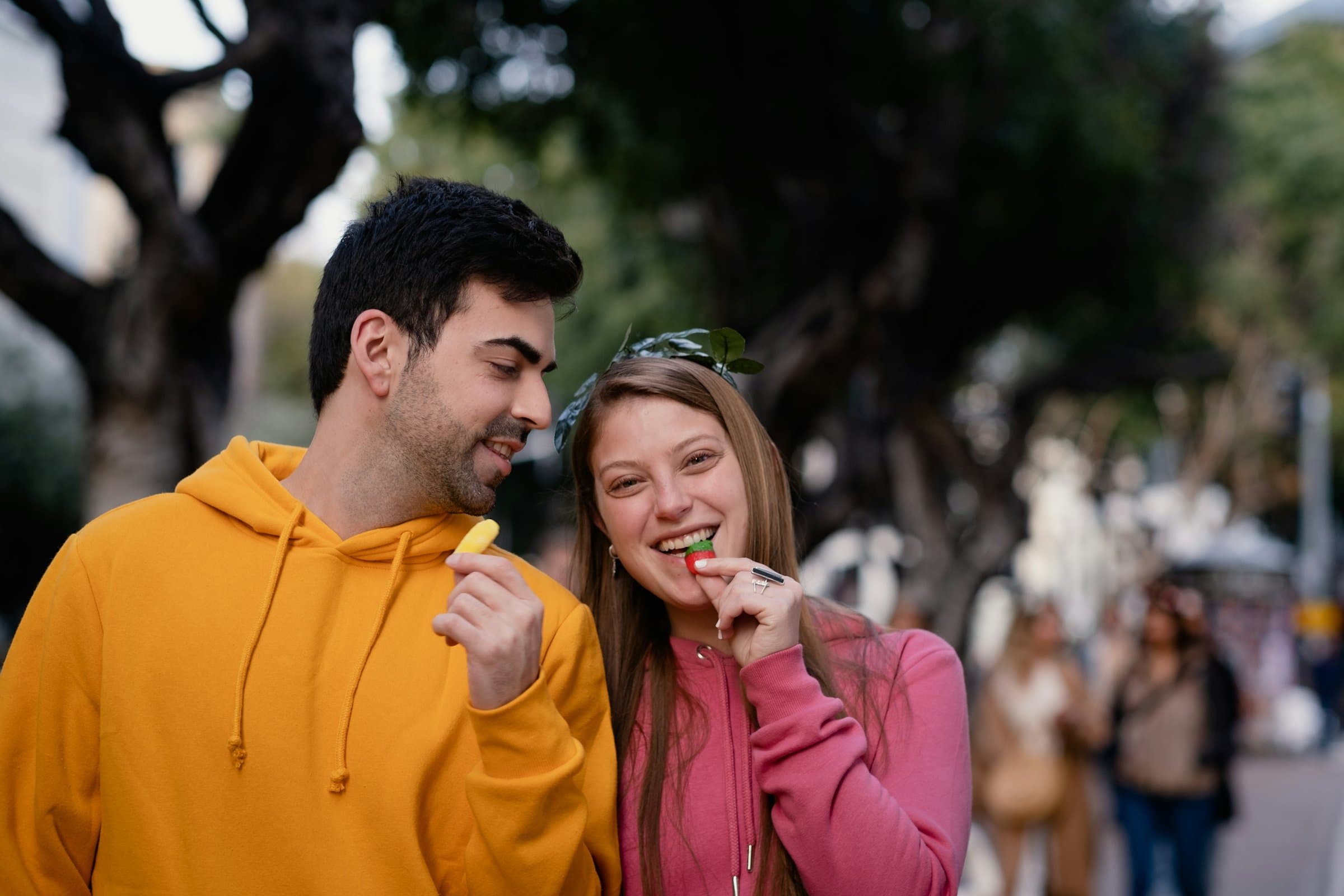 A man and a woman sharing lollies and grinning as they stroll down a tree-lined street