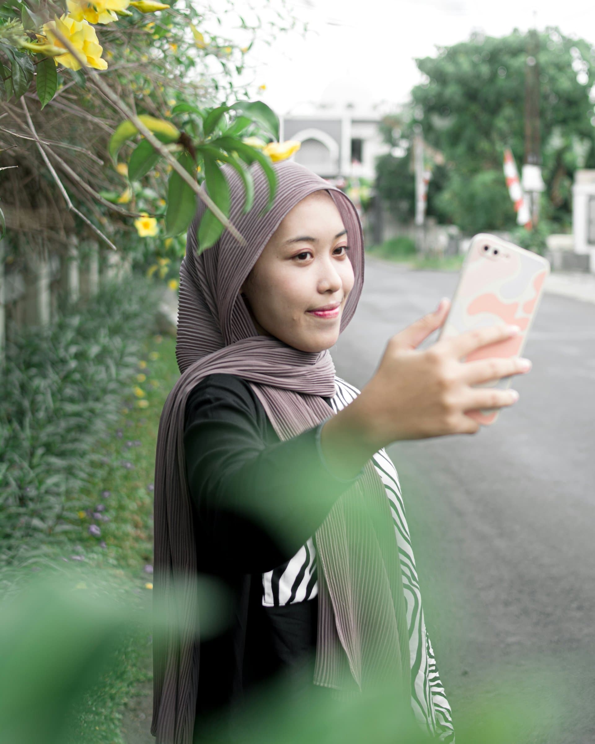 A young woman in a mauve hijab smiling softly as she takes a selfie on a leafy street