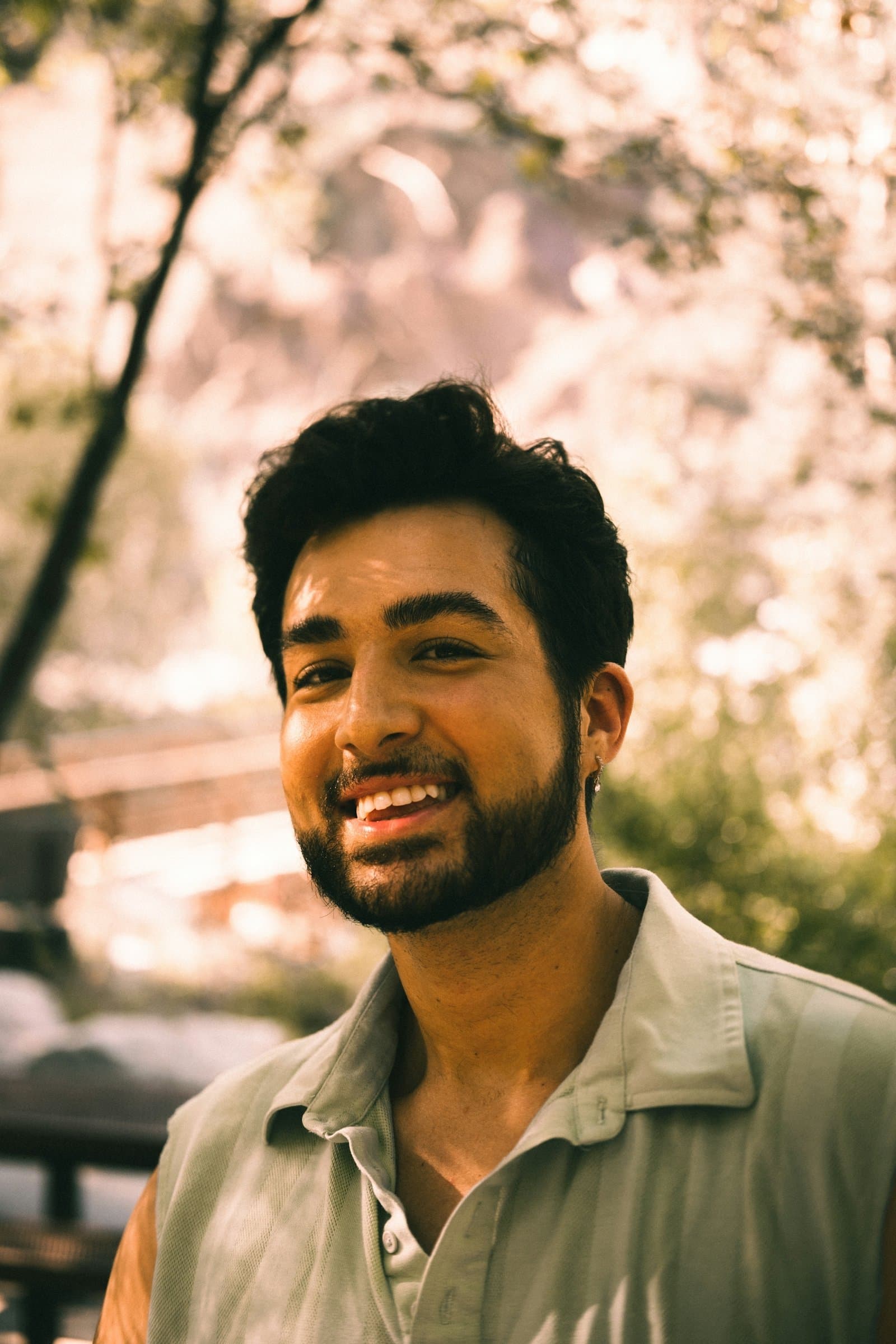 A bearded South Asian man in a sage shirt smiles warmly at the camera in soft light under trees.